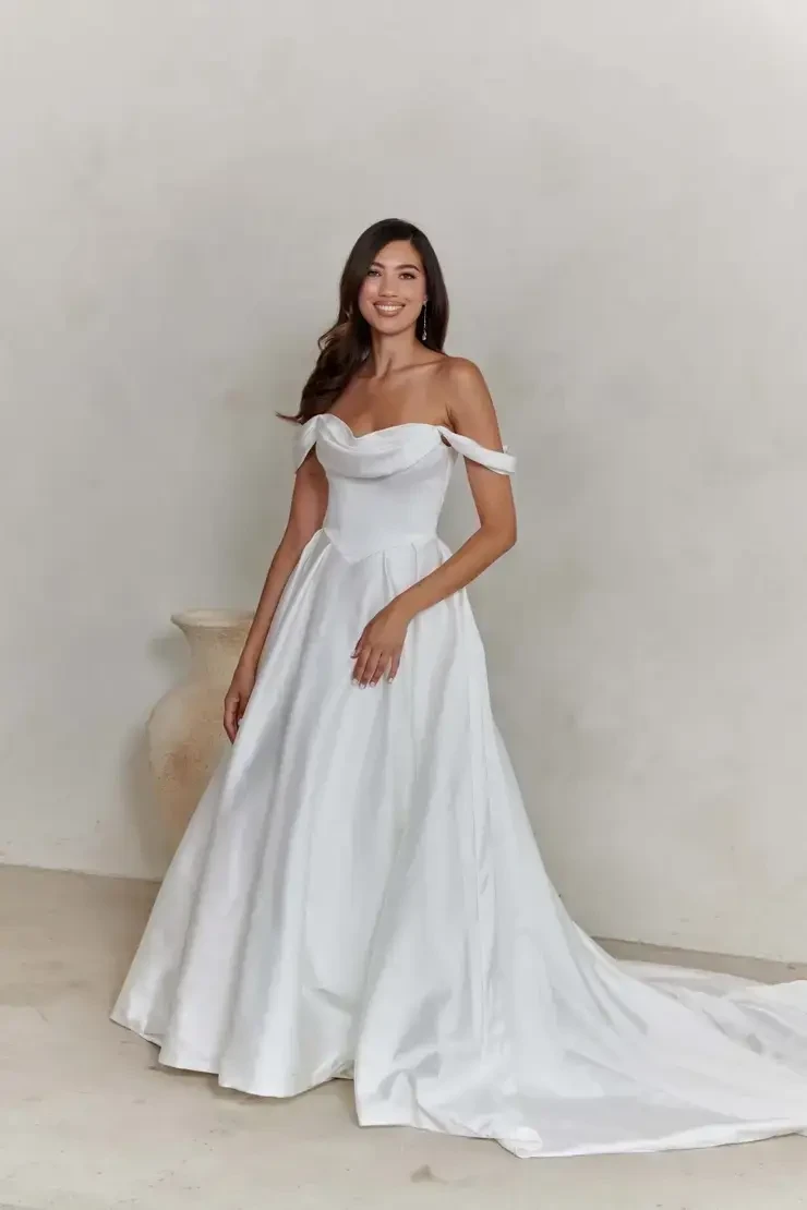 A smiling woman in an elegant, off-shoulder white wedding gown with a flowing train. She stands in a minimalist beige room with a large ceramic vase.