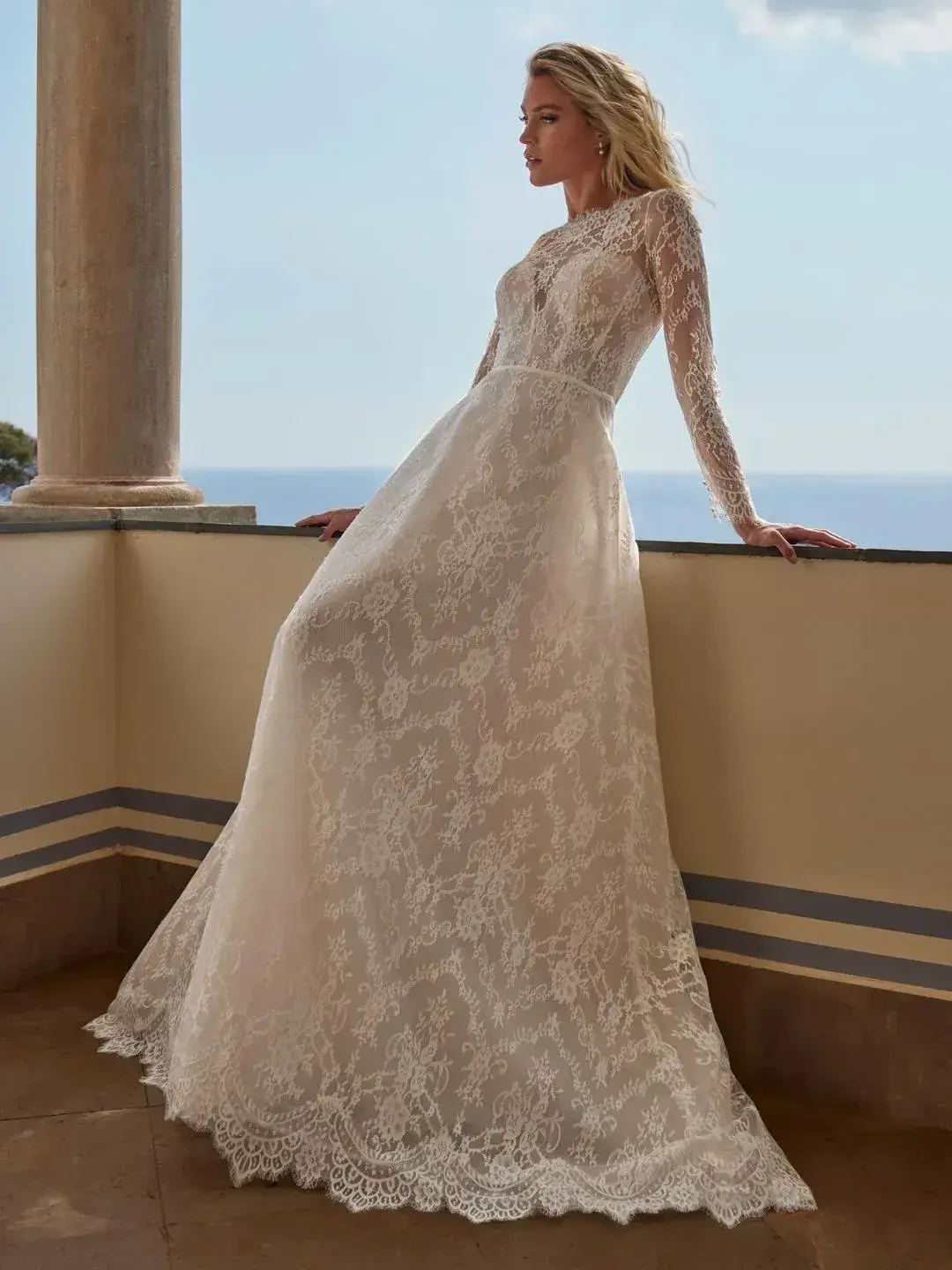 A woman wearing a long-sleeved lace wedding dress, standing on a balcony overlooking the sea.