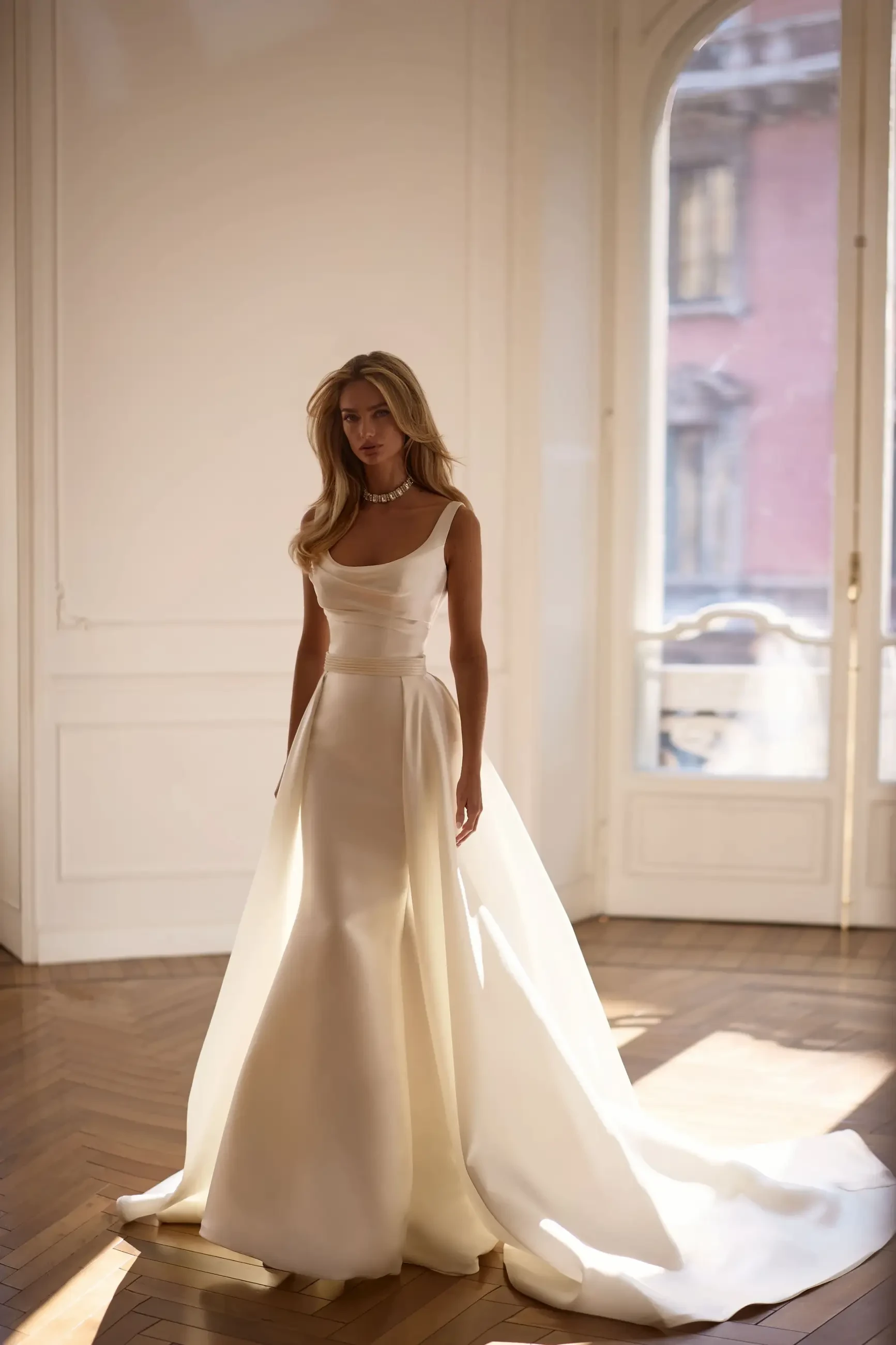 A woman in a flowing white dress stands elegantly in a well-lit room with large windows.