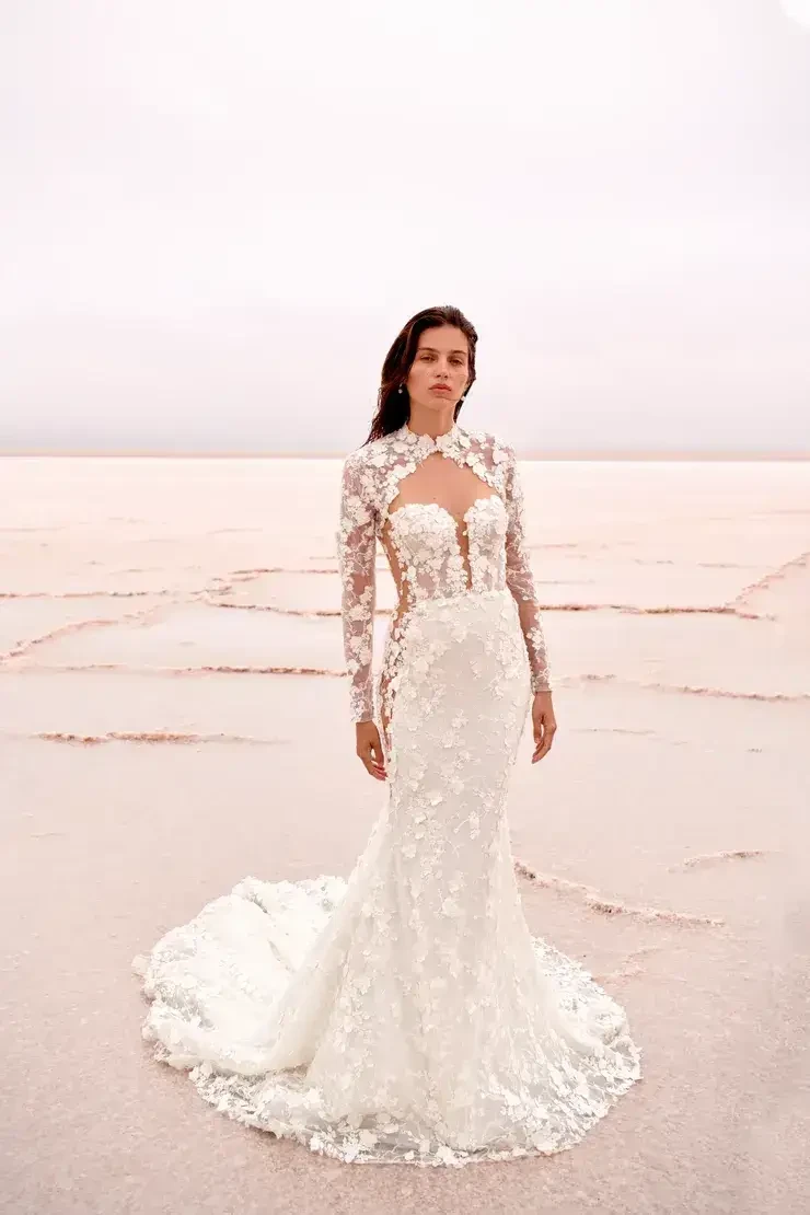A woman stands on a vast, pink salt flat, wearing an elegant white lace wedding dress with floral details and a trailing hem, exuding serenity and grace.