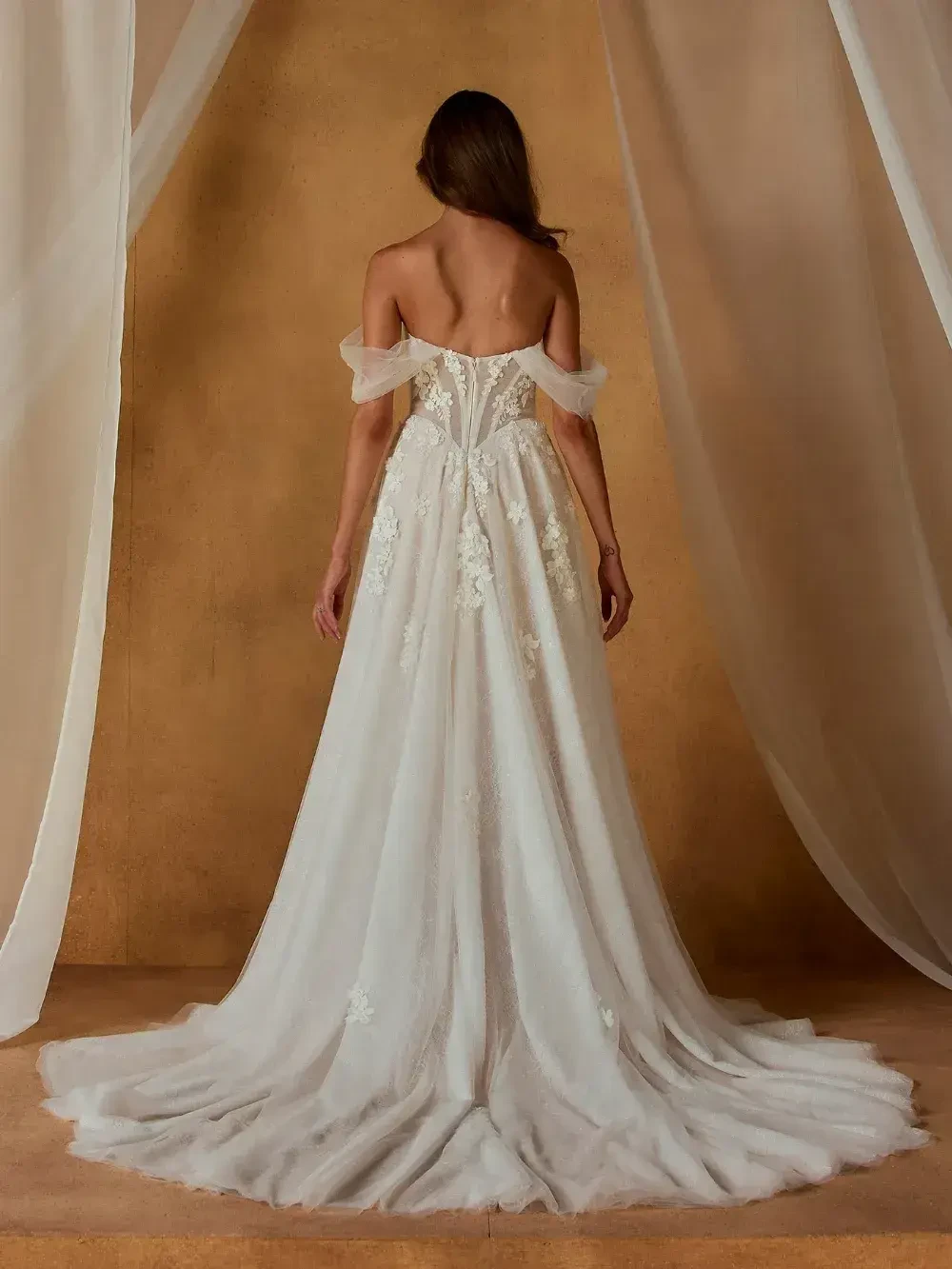 A woman in a wedding dress with off-the-shoulder sleeves, viewed from the back, standing against a textured background. The dress is long and flowing with intricate detailing.