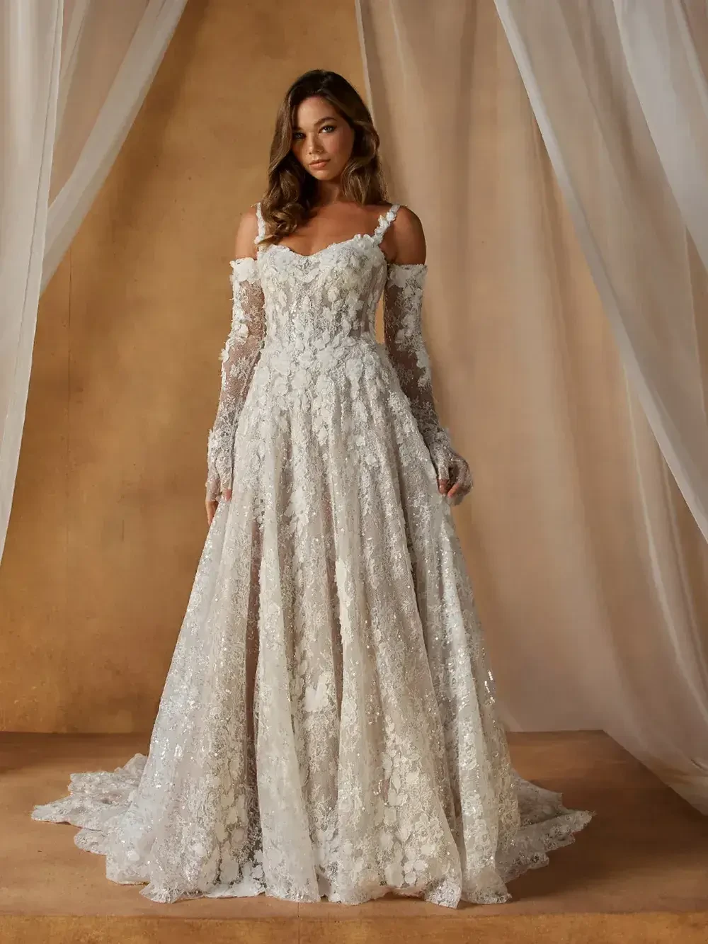 A woman in a stunning white wedding dress with intricate lace detailing and long lace sleeves, standing elegantly against a softly draped backdrop.