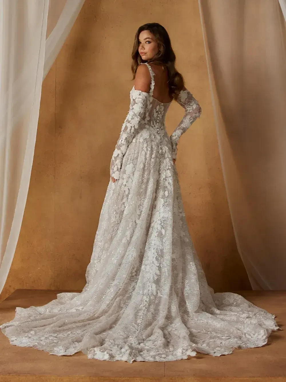 A woman in a long, lace wedding dress stands with her back to the camera, showcasing the intricate details of the gown.