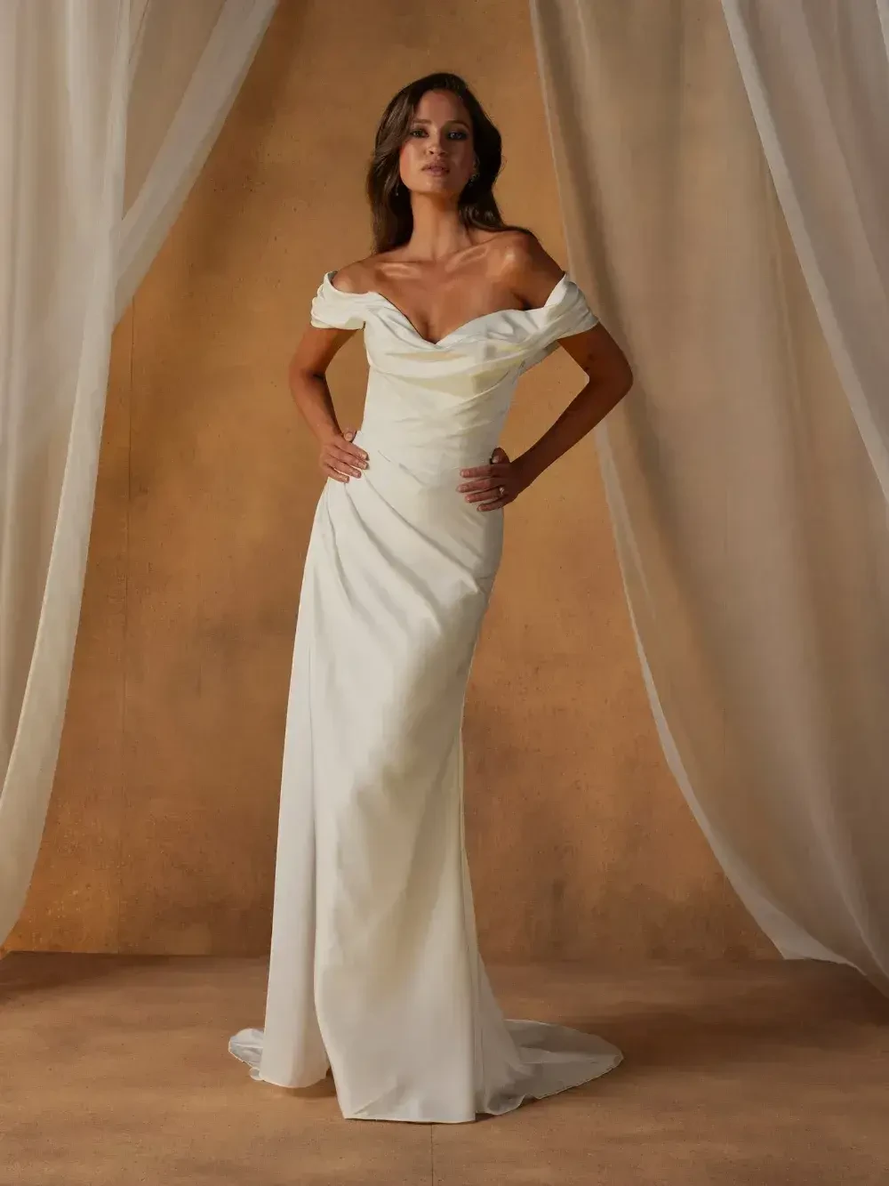 A woman modeling an off-the-shoulder white gown against a soft, neutral backdrop.