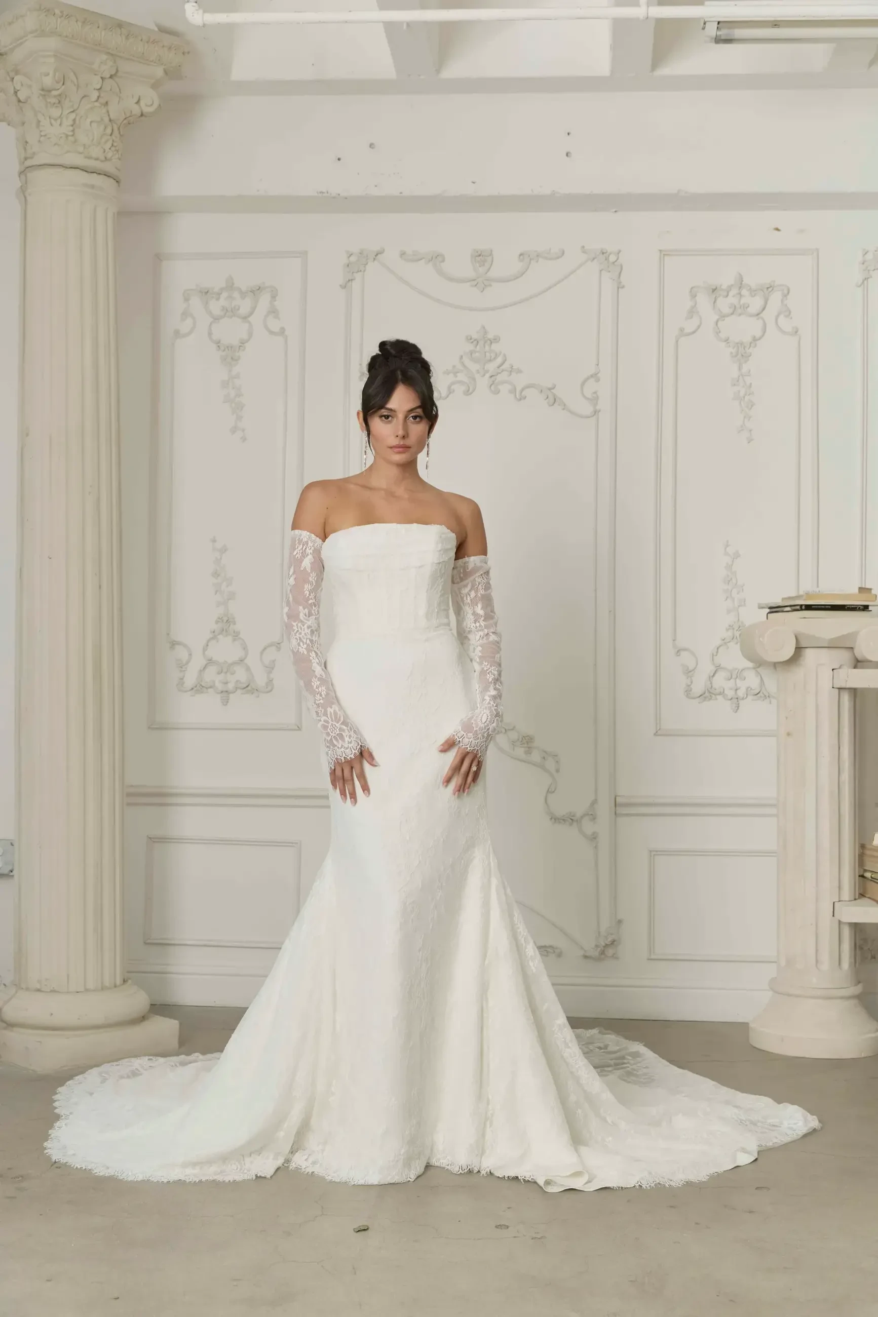 A model stands in a fitted white wedding gown with long lace sleeves against a decorative wall and columns.