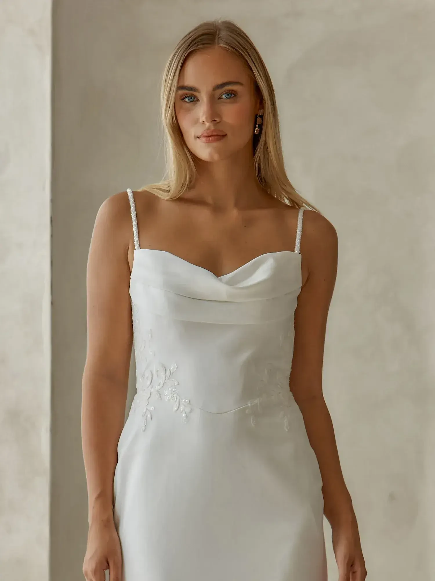 A young woman wearing a simple, elegant white dress with thin straps, standing against a neutral-toned background.