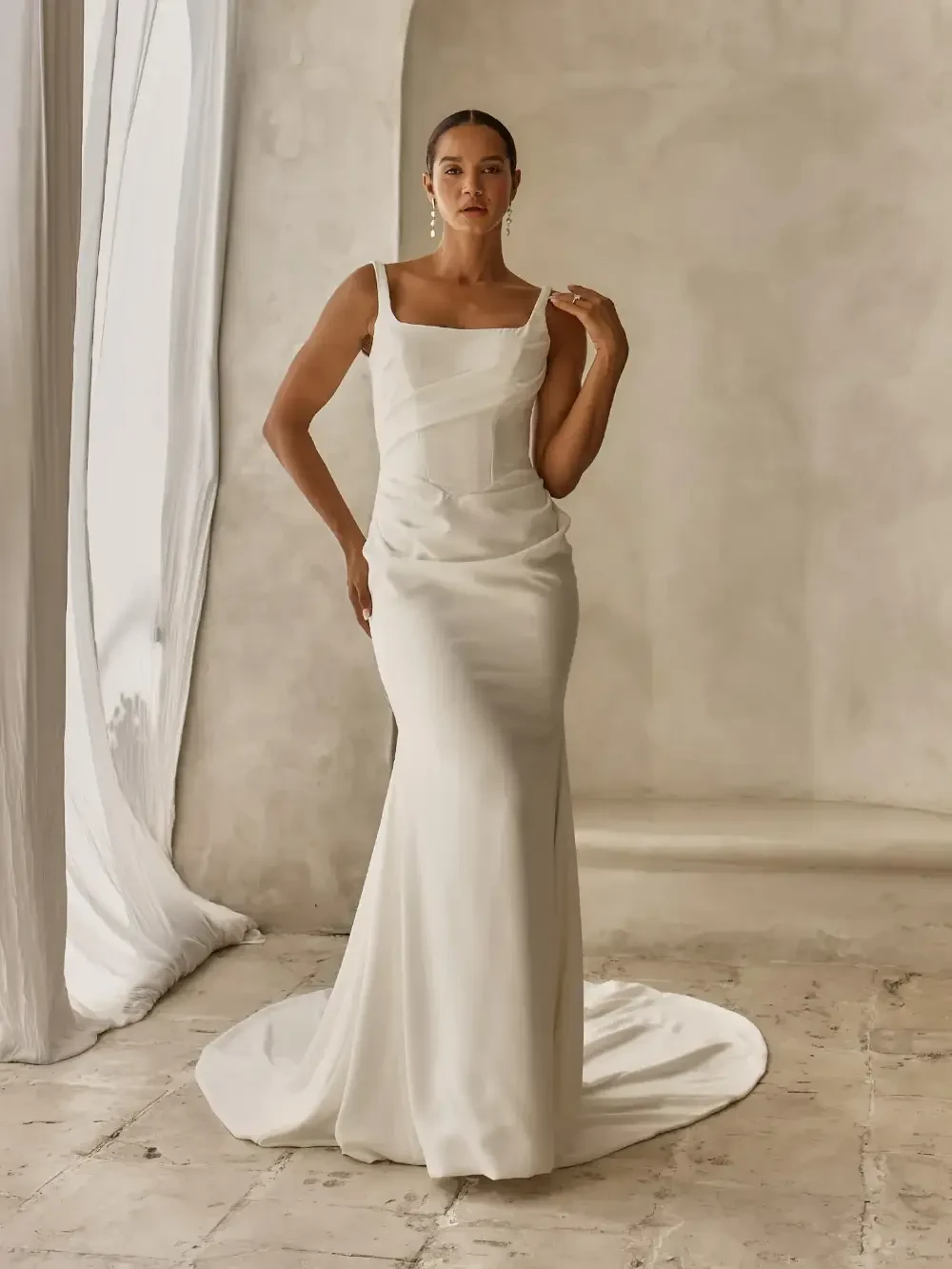A model wearing a sleek, elegant white dress with a train, posing in a softly lit environment with a minimalistic background.
