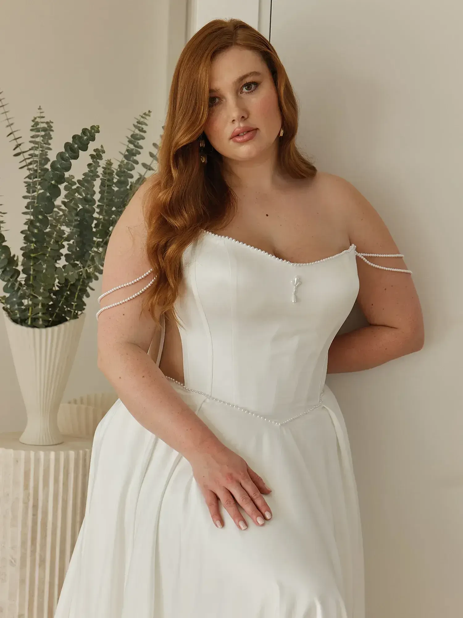 A person wearing a white dress with off-the-shoulder straps, standing next to a decorative plant in a vase. Their hair is styled in loose waves.