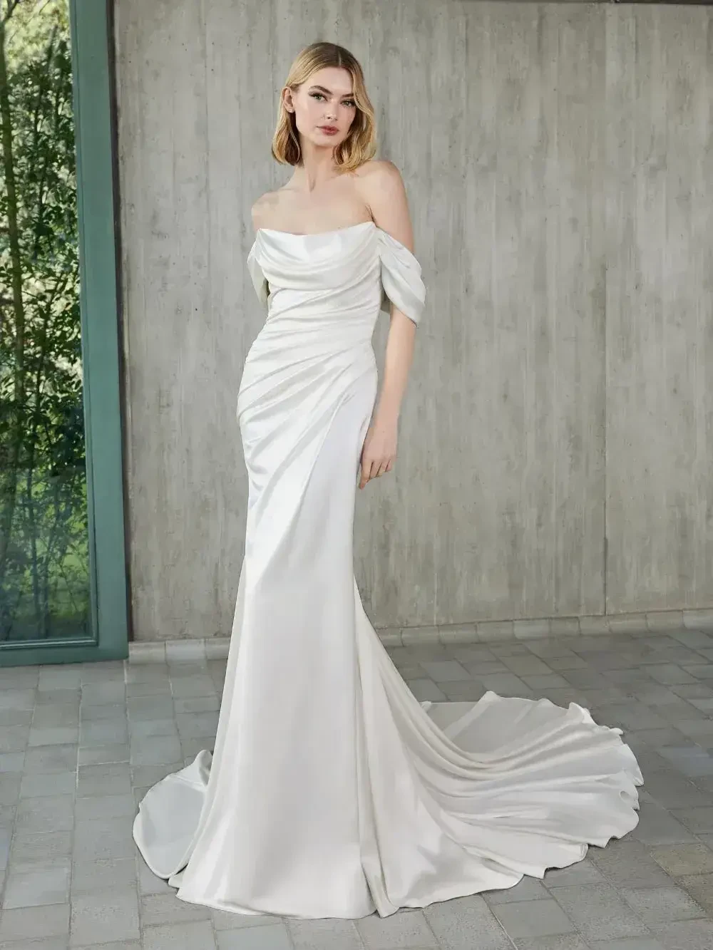 A woman wearing an elegant off-the-shoulder white wedding gown, standing in front of a modern textured wall with greenery in the background.